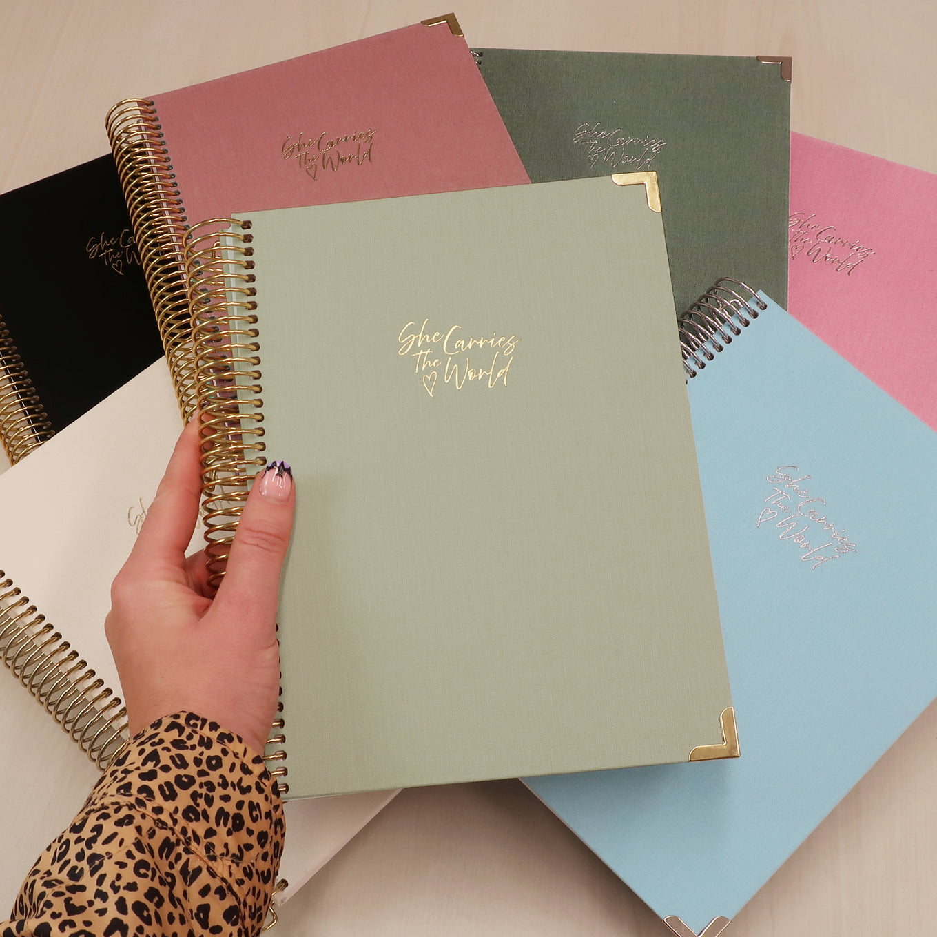 Collection of linen pregnancy journals on a wooden surface with a hand holding one. 'She Carries The World' logo embossed in metallics 