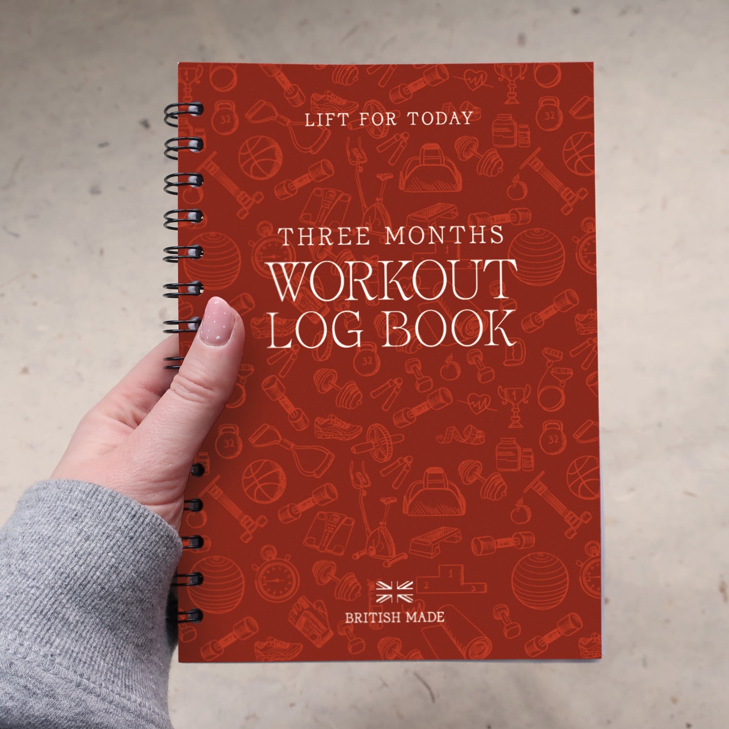 A hand holding a orange spiral-bound workout log book with the title 'Three Months Workout Log Book' on the cover, photographed against a light textured floor.