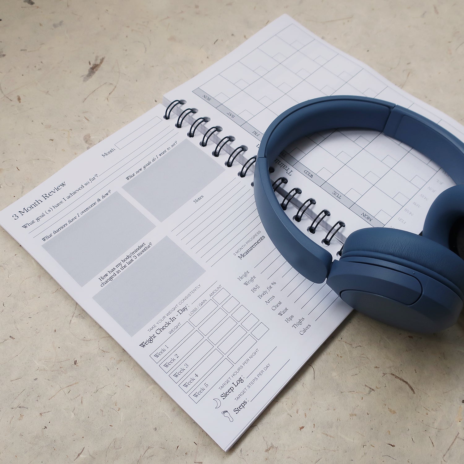 Blue headphones on a workout log book open on the 3 month review page. With boxes to fill in like sleep log, steps, and body measurements. 