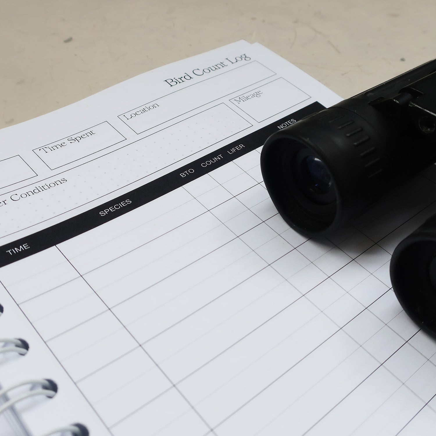 wayzgoose Bird Watching Log Book Open On The Bird Count Log Page, Laid On A Textured Background With Black Binoculars 